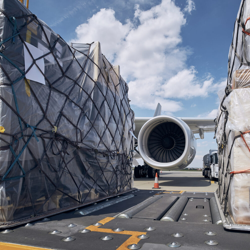 Air freight product sitting on runway in preparation before a flight with cargo containers and air plane jet engine in background.